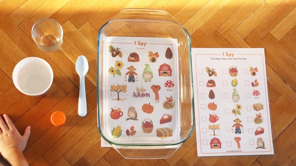 Activity setup on a tray: A Montessori activity tray holding an "I Spy: Autumn" printable sheet, a checklist, a dry-erase marker, and a clear plastic cup of tinted water.