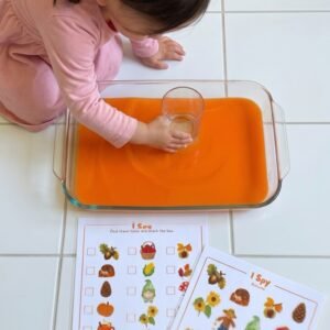 A Montessori activity tray holding an "I Spy: Autumn" printable sheet, a checklist, a dry-erase marker, and a clear plastic cup of tinted water.